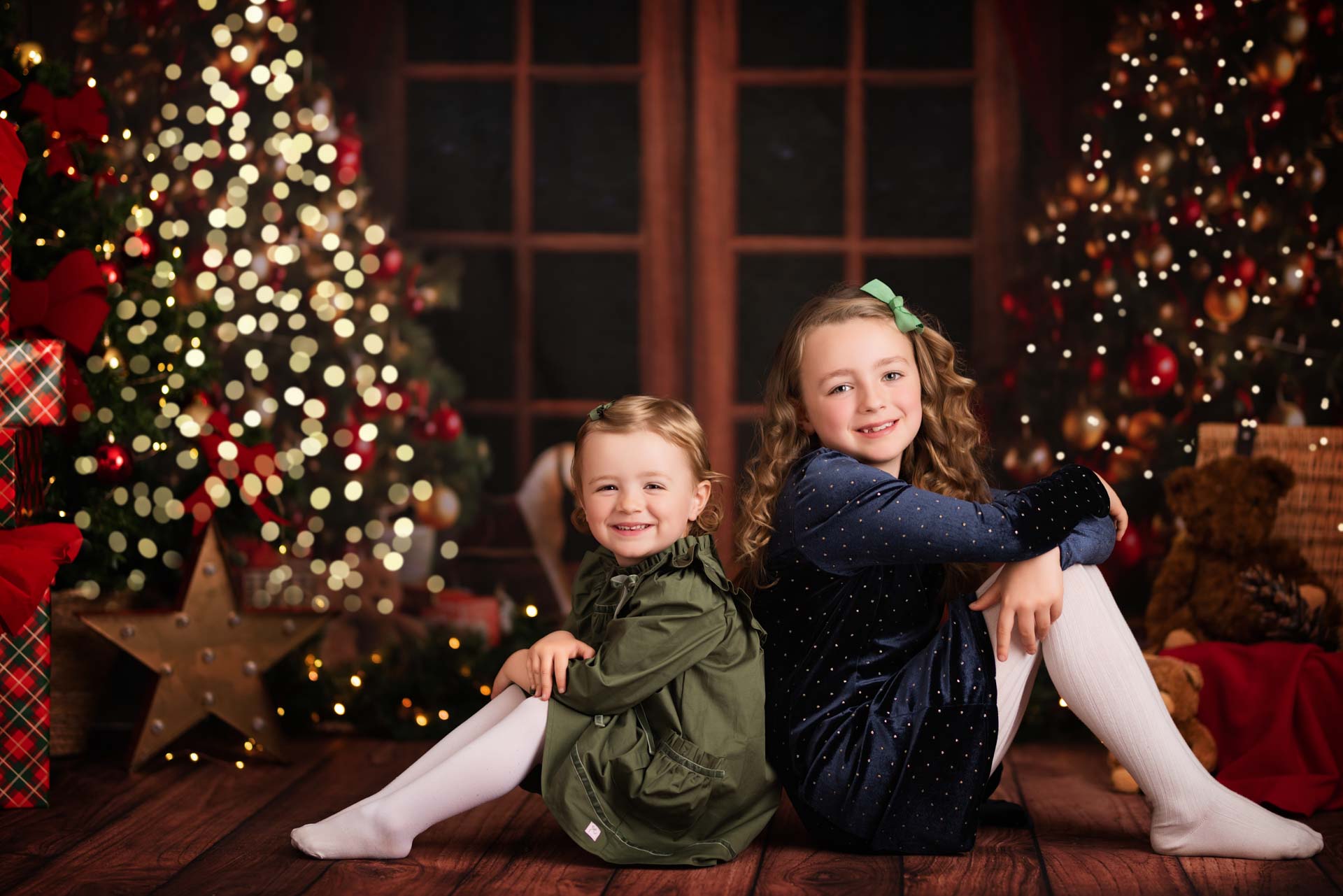 Christmas Photo Shoot Oxfordshire Two sisters sit back to back. One is wearing a blue dress, the other green. They both have white tights and green bows in their hair. The background is green and red with two Christmas trees.