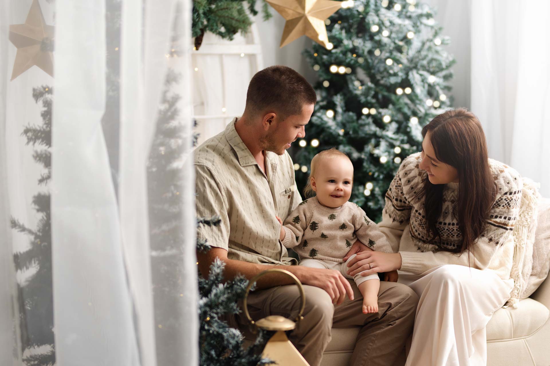 Family Christmas Photos Cotswolds Mother and father looking down at the son adoringly. They are sat by two large green and white Christmas trees. Golden stars hang down from the ceiling.