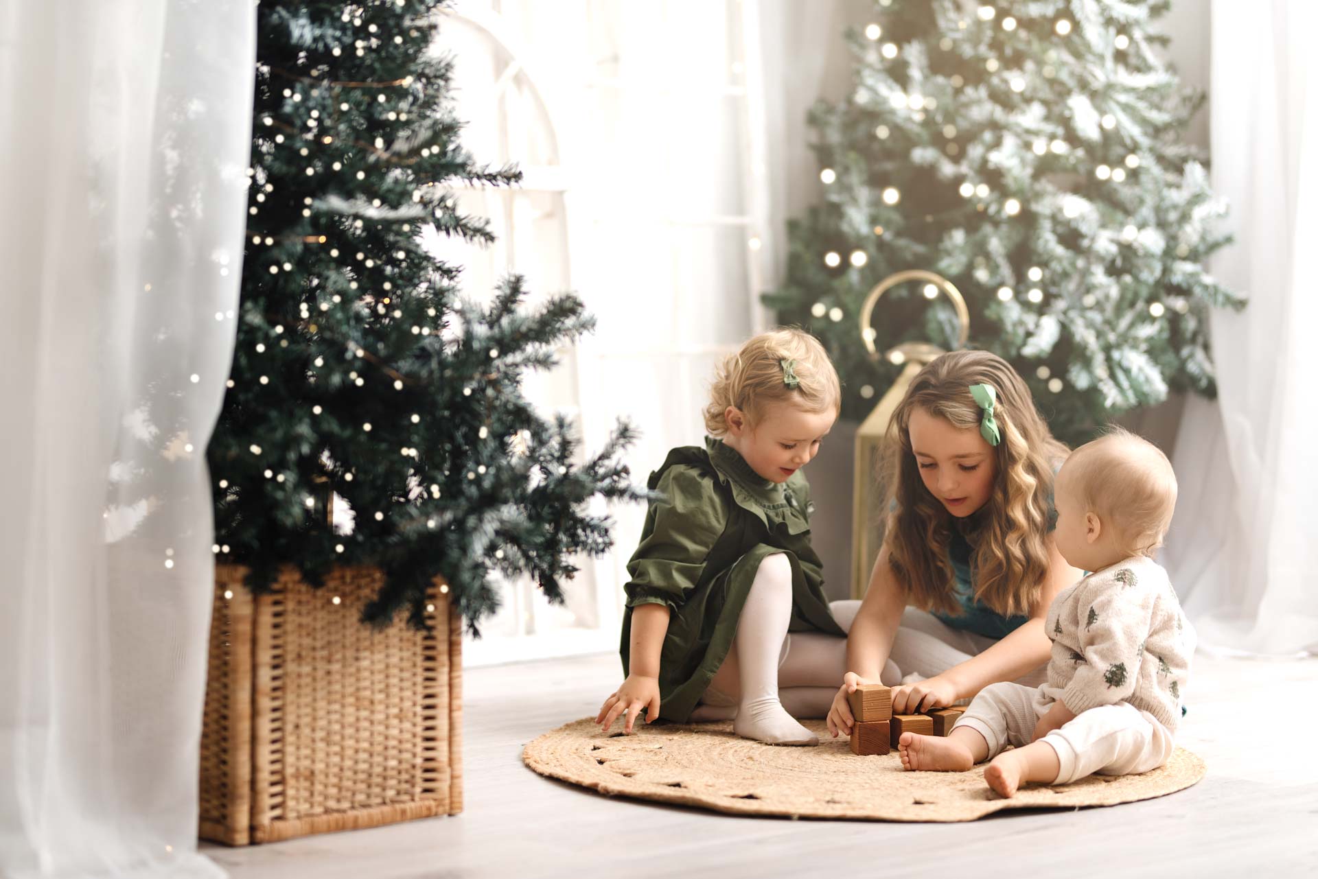 Kids Christmas Photoshoot Oxfordshire Three young children sitting on a mat playing with wooden blocks. In the background are two Christmas trees with twinkly lights.
