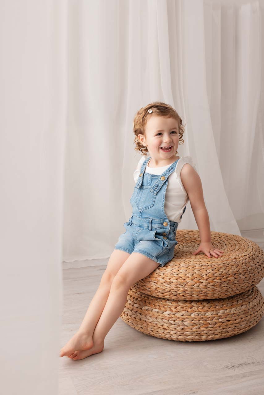Nursery Photography Cotswolds Girl wearing a white t-shirt and denim dungarees smiles during her nursery photoshoot