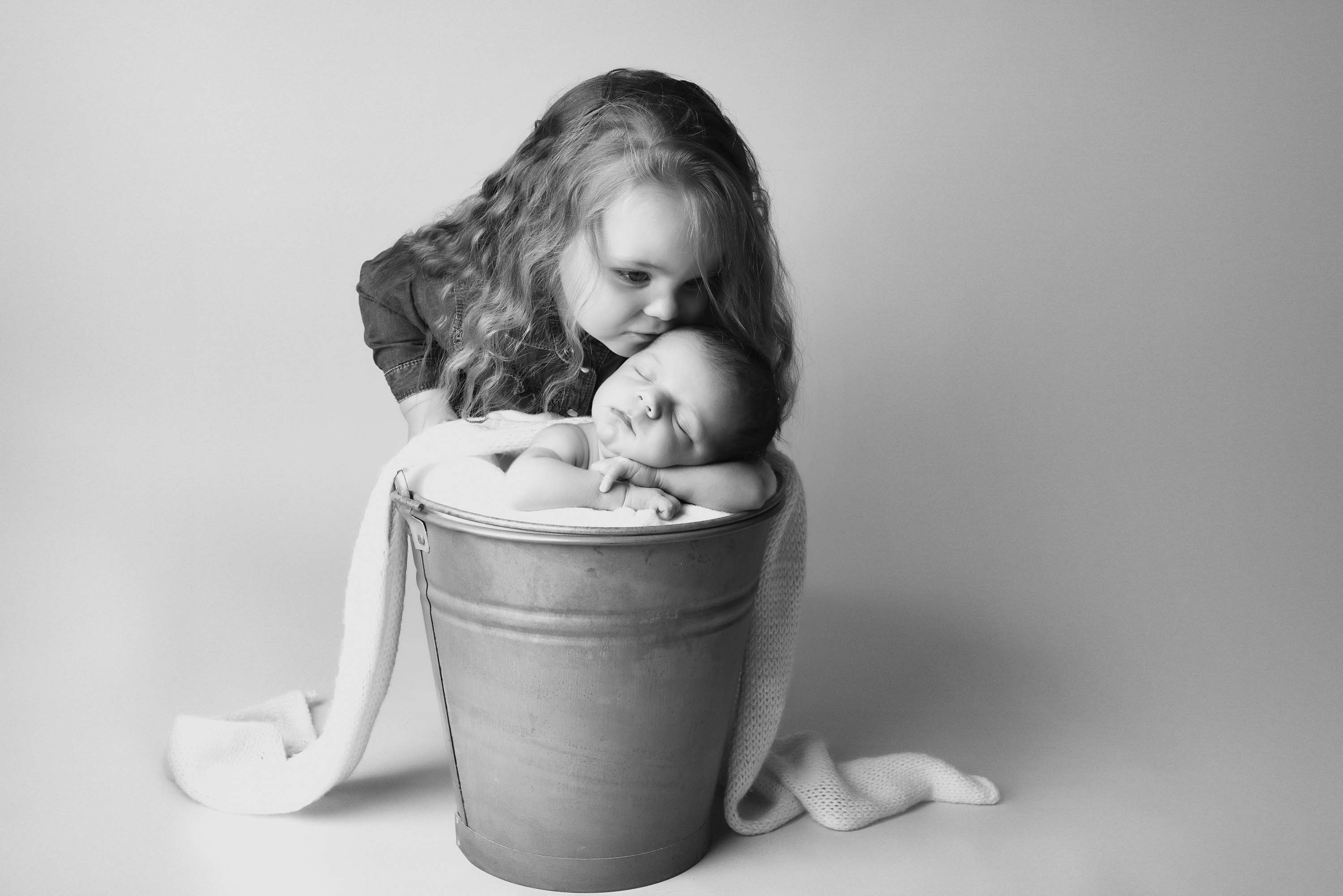 Two year old girl kissing her sibling on the forehead. Her brother is sleeping in a bucket.