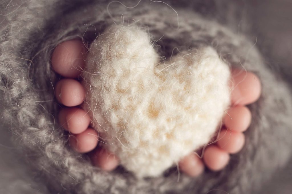 Ten beautiful baby toes holding a knitted white heart surrounded by grey woolly blanket