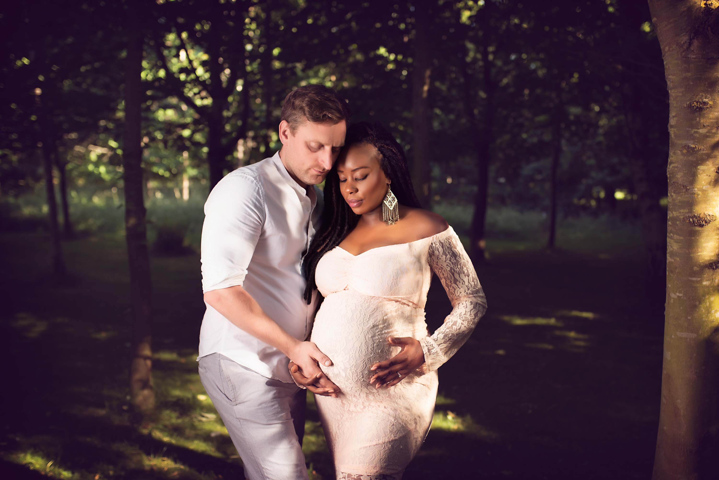 Pregnant couple standing under trees looking at their baby bump