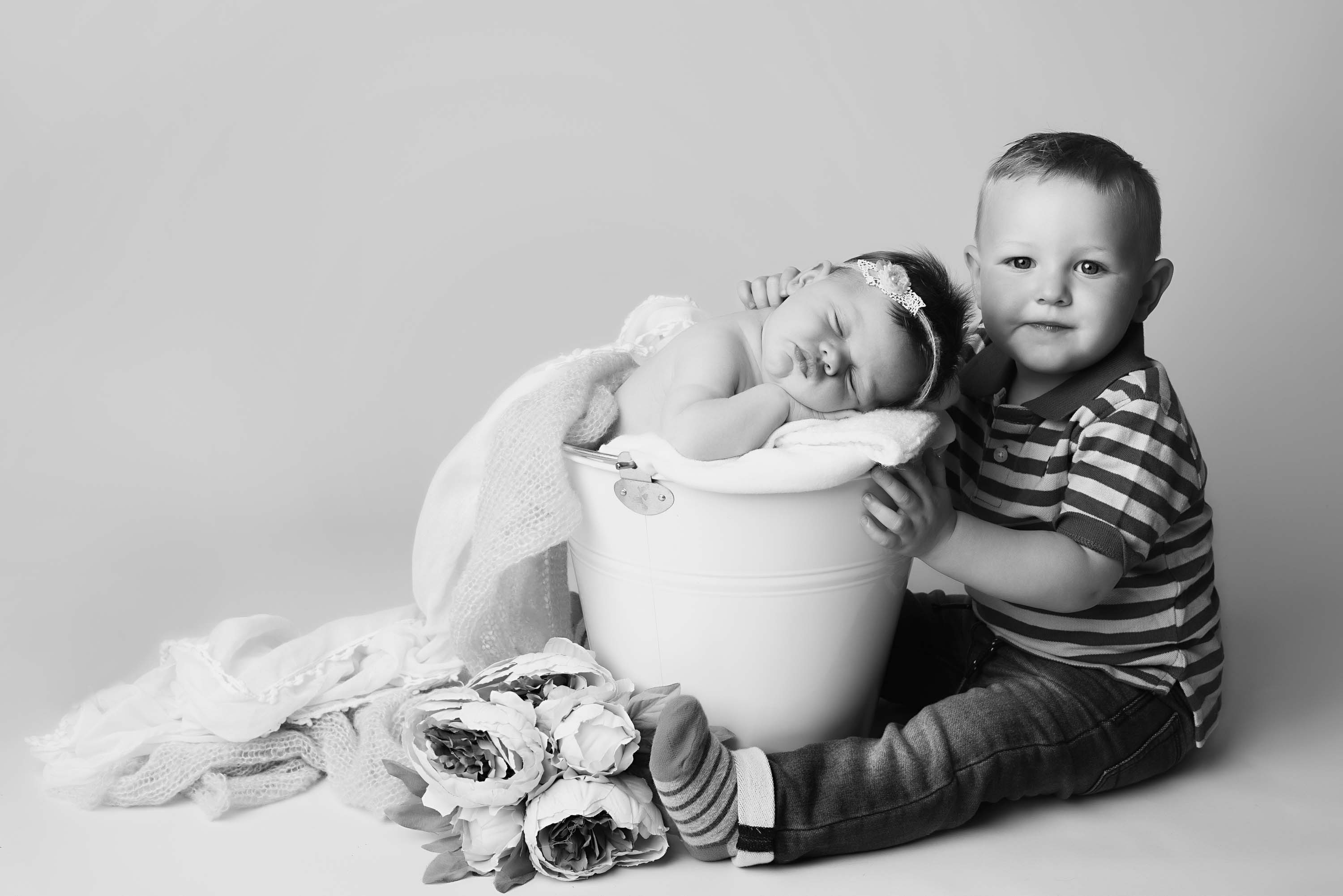 Photo of siblings. Brother sitting next to baby girl who is posed in a bucket