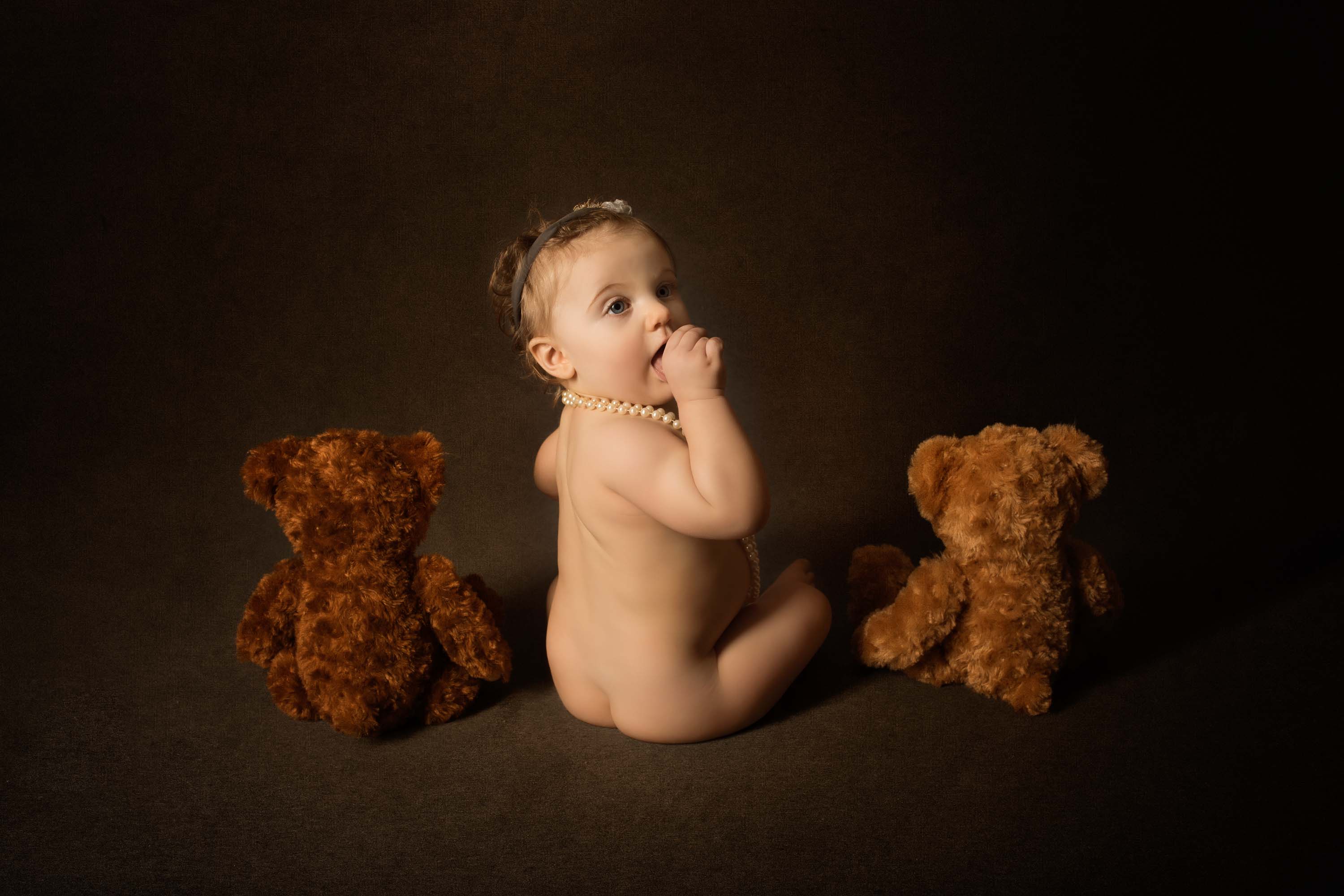 One year old sitting facing away from the camera with thumb in her mouth and a teddy bear on either side