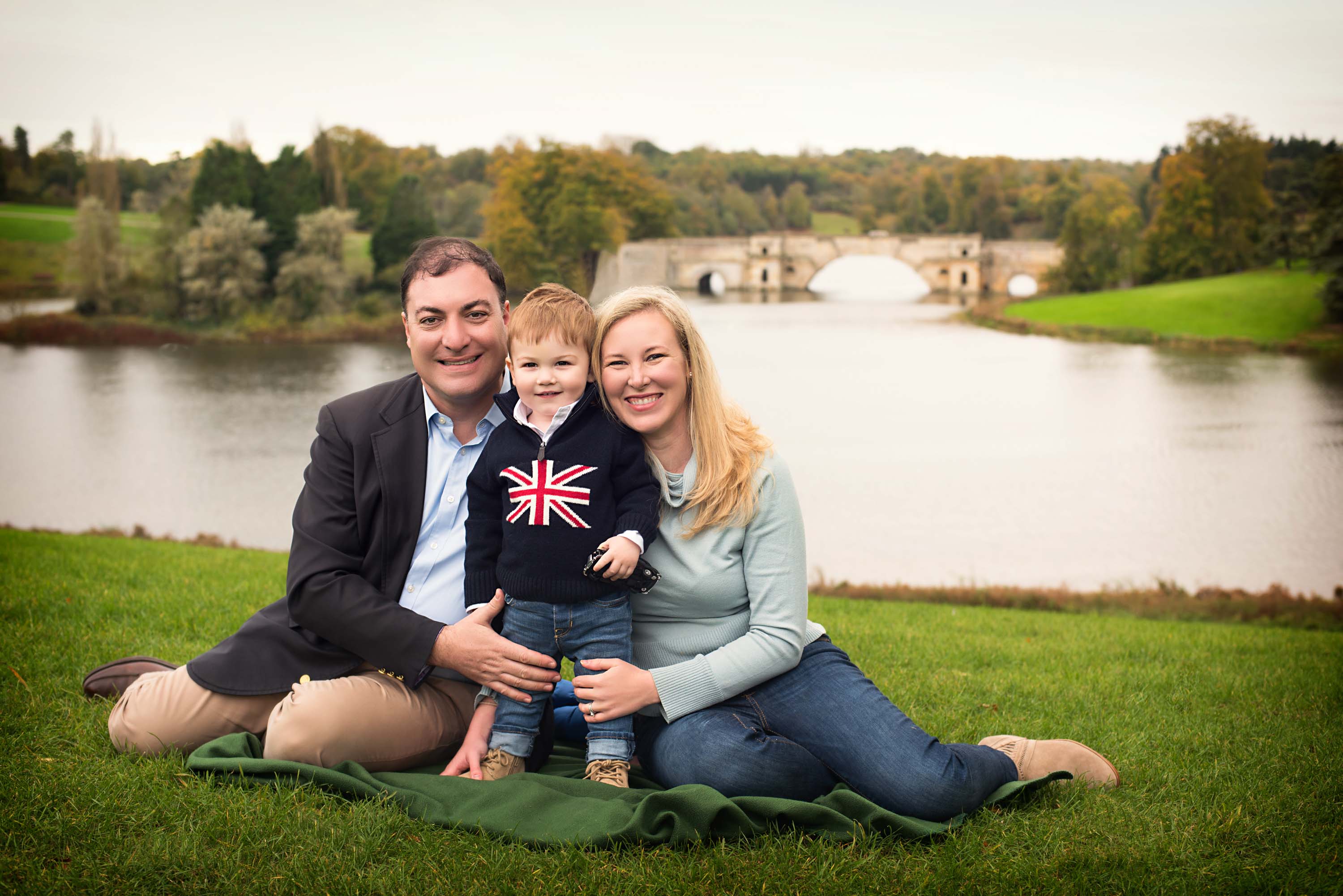 Family of three sitting on blanket at Blenheim Palace with lake and bridge in background