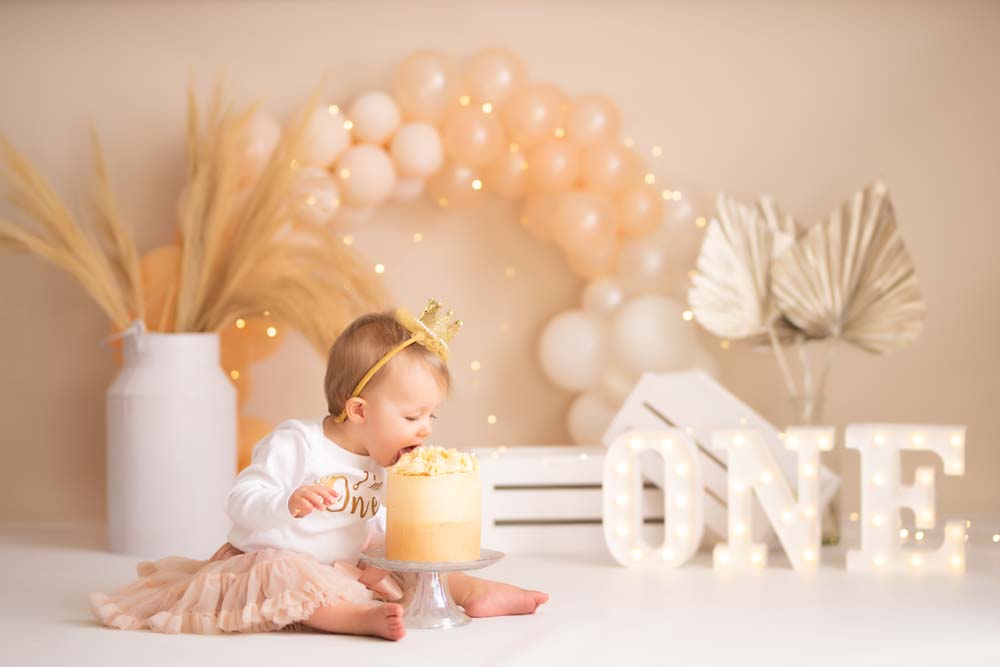 Girl wearing a tutu sticking her face in her birthday cake at her photoshoot in front of white and peach balloons