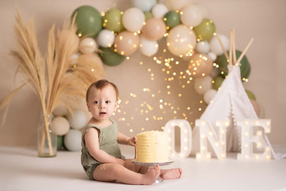 Boy sitting next to cake at his first birthday photoshoot in front of green and cream balloons