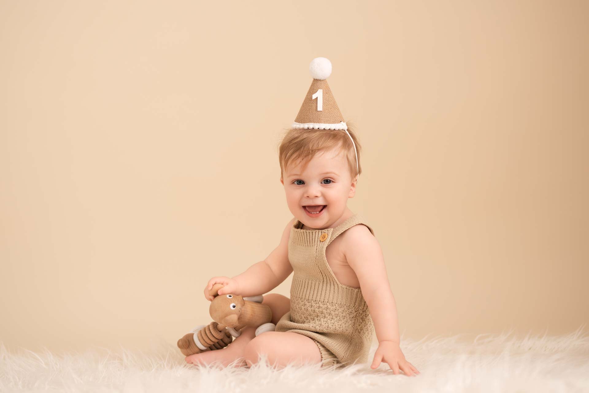 Smiling baby boy sitting on a white blanket. He is wearing a light brown romper and party hat with the number one on it.