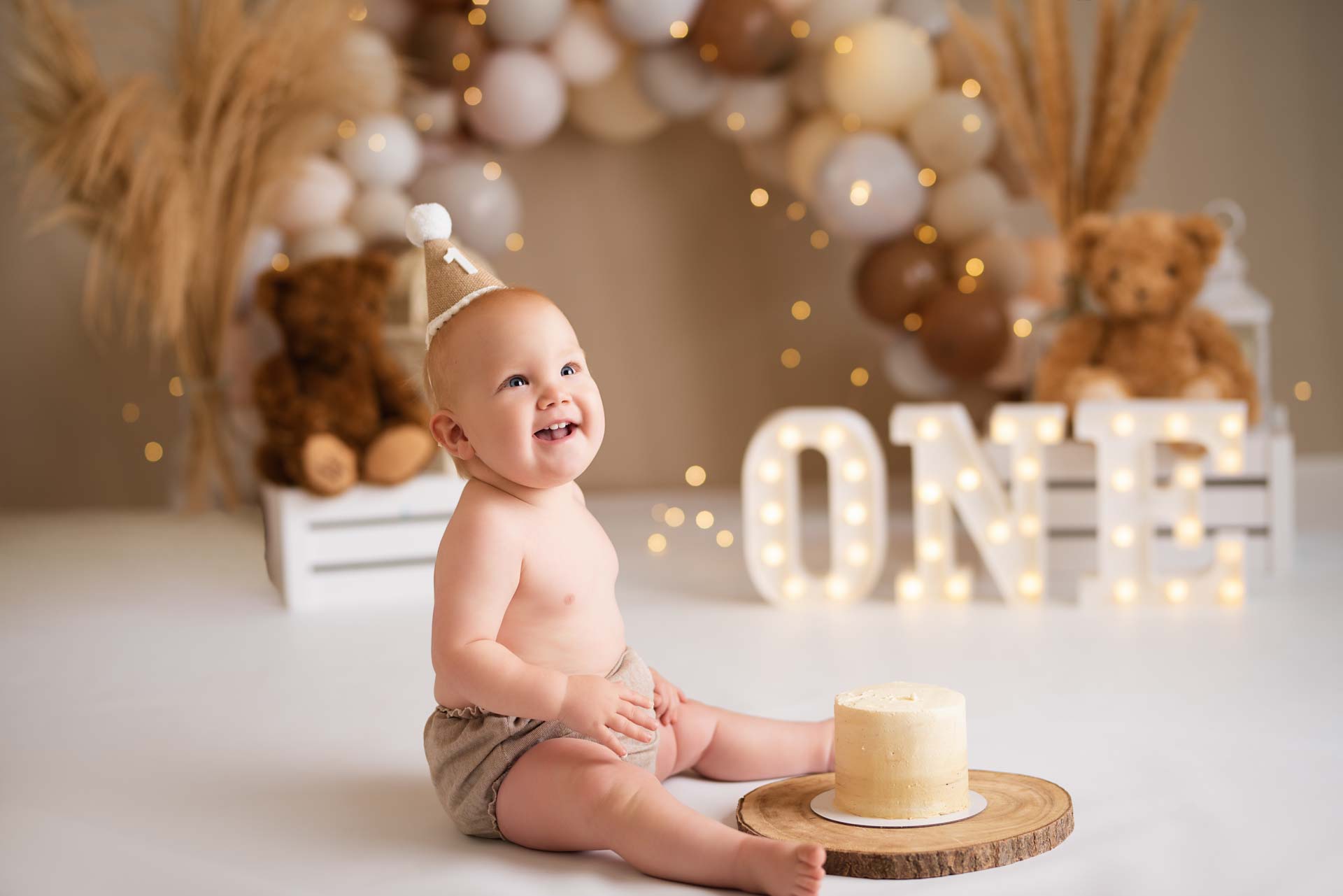 One year old boy sitting next to his birthday cake. The cake is on a wooden board. In the background are brown and white balloons and teddy bears