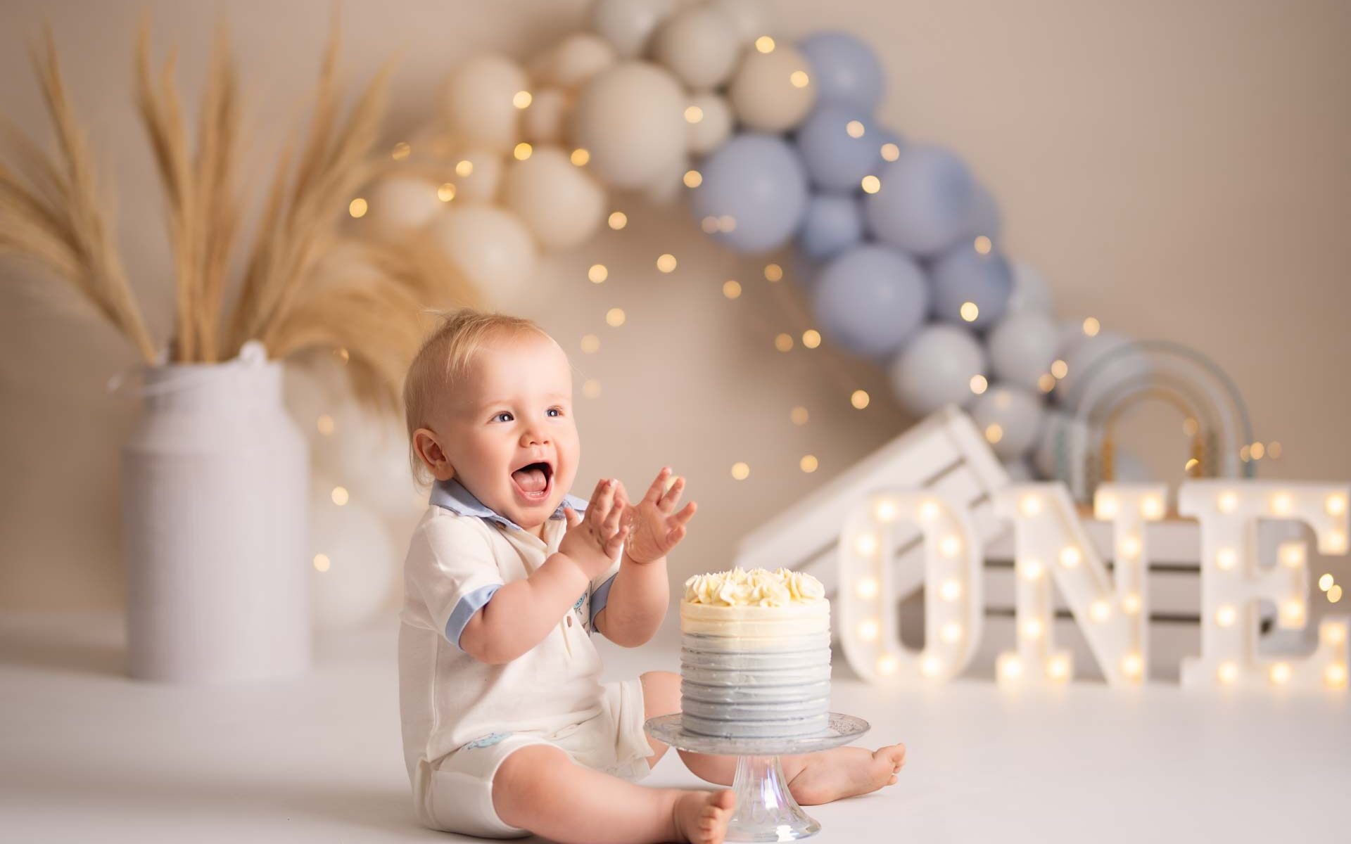 Little boy sitting next to his blue and white birthday cake. He is clapping and clearly excited to eat his cake
