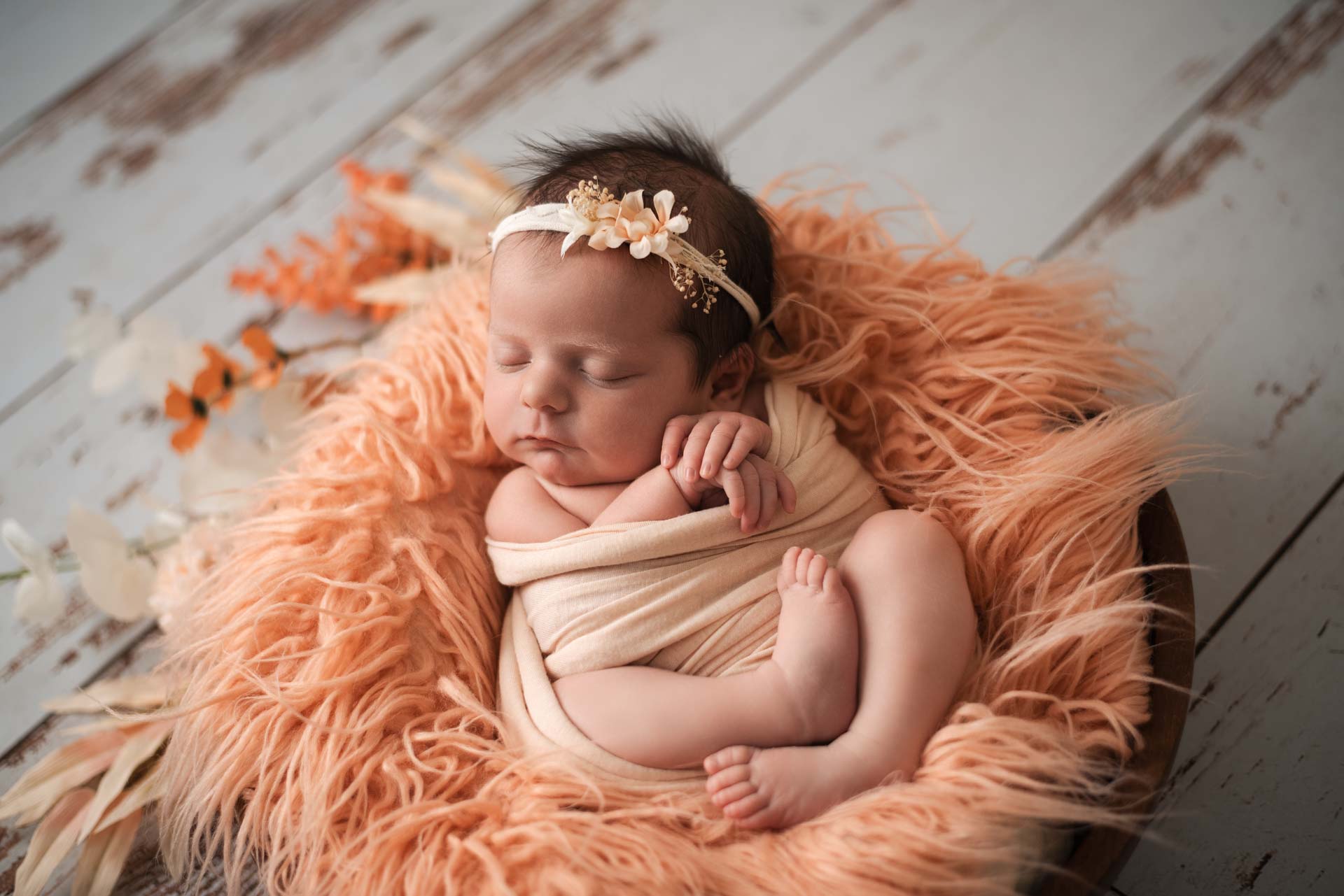 Newborn baby wrapped in a peach coloured blanket resting in a peach coloured fluffy blanket in a wooden bowl