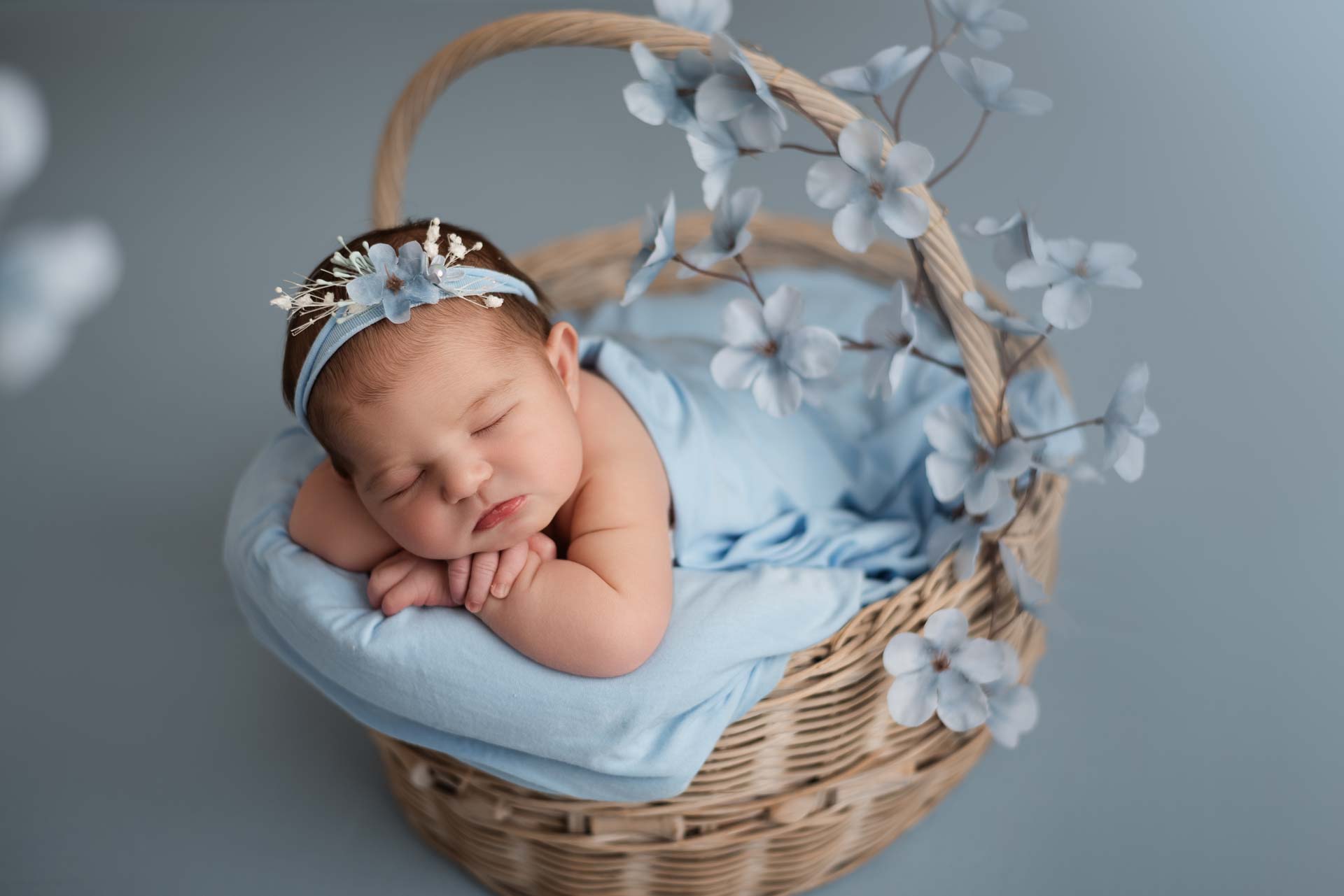 Baby girl lying on her front posed in a wicker basket. There is a blue blanket, headband and flowers