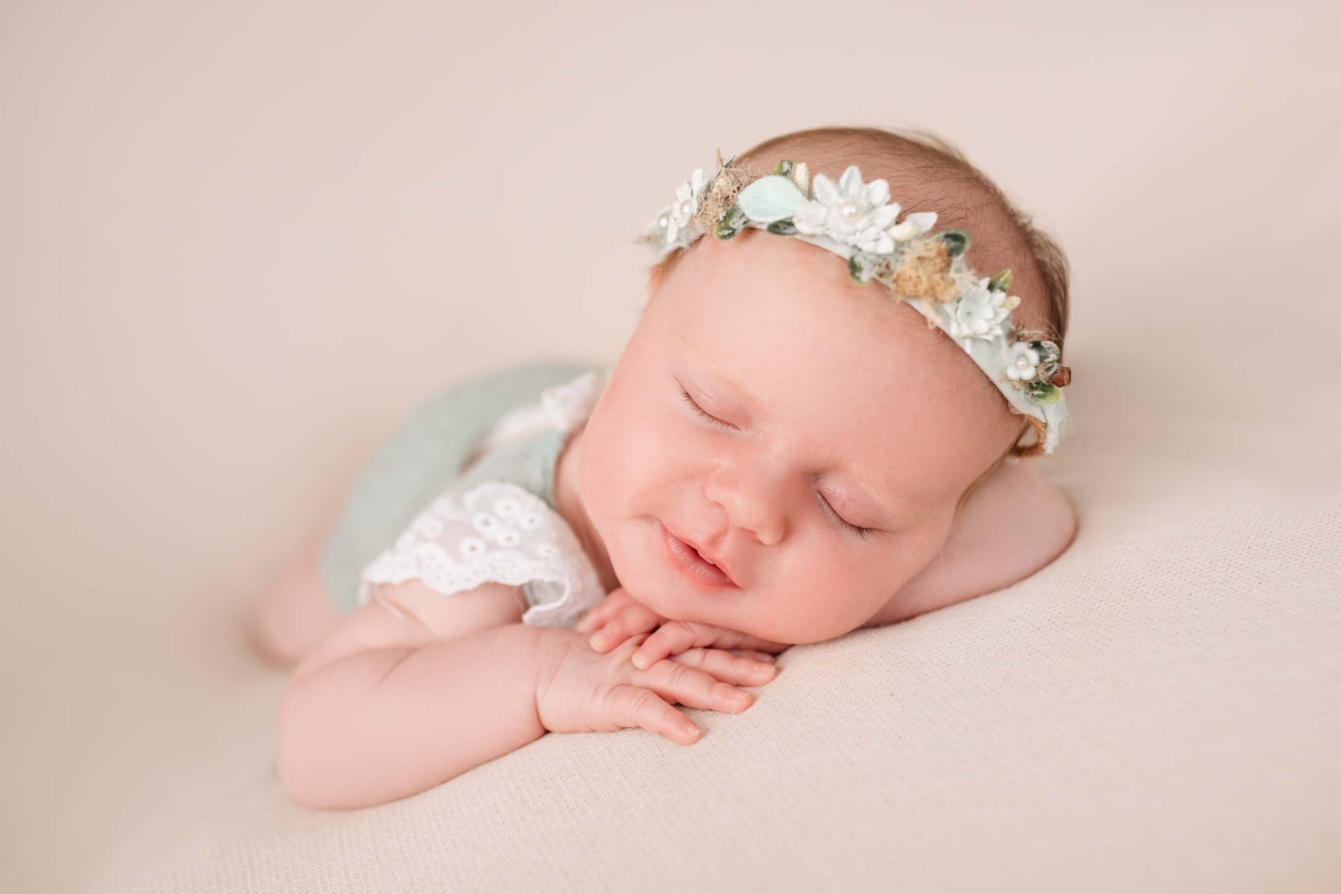 Newborn baby girl wearing floral headband and lacey romper. They are both white and pale green.