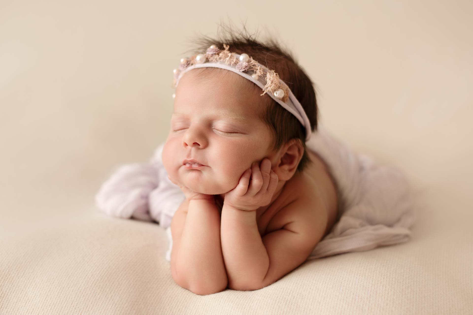 A composite photo of a newborn baby with her hands resting on her arms. She is wearing a floral lilac headband with pearls in it.