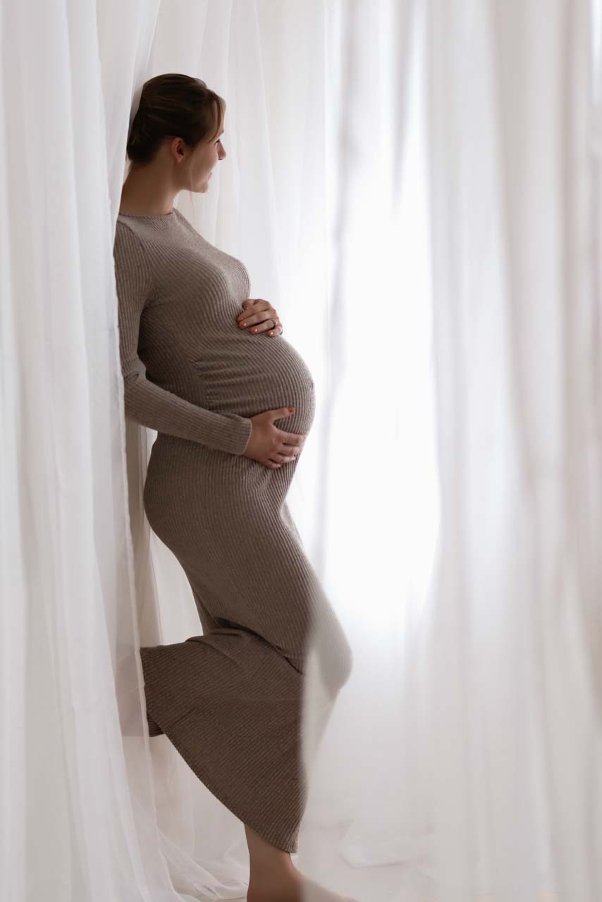 Expectant mother in a stretchy long beige dress holds her bump and looks out of the window dreaming.