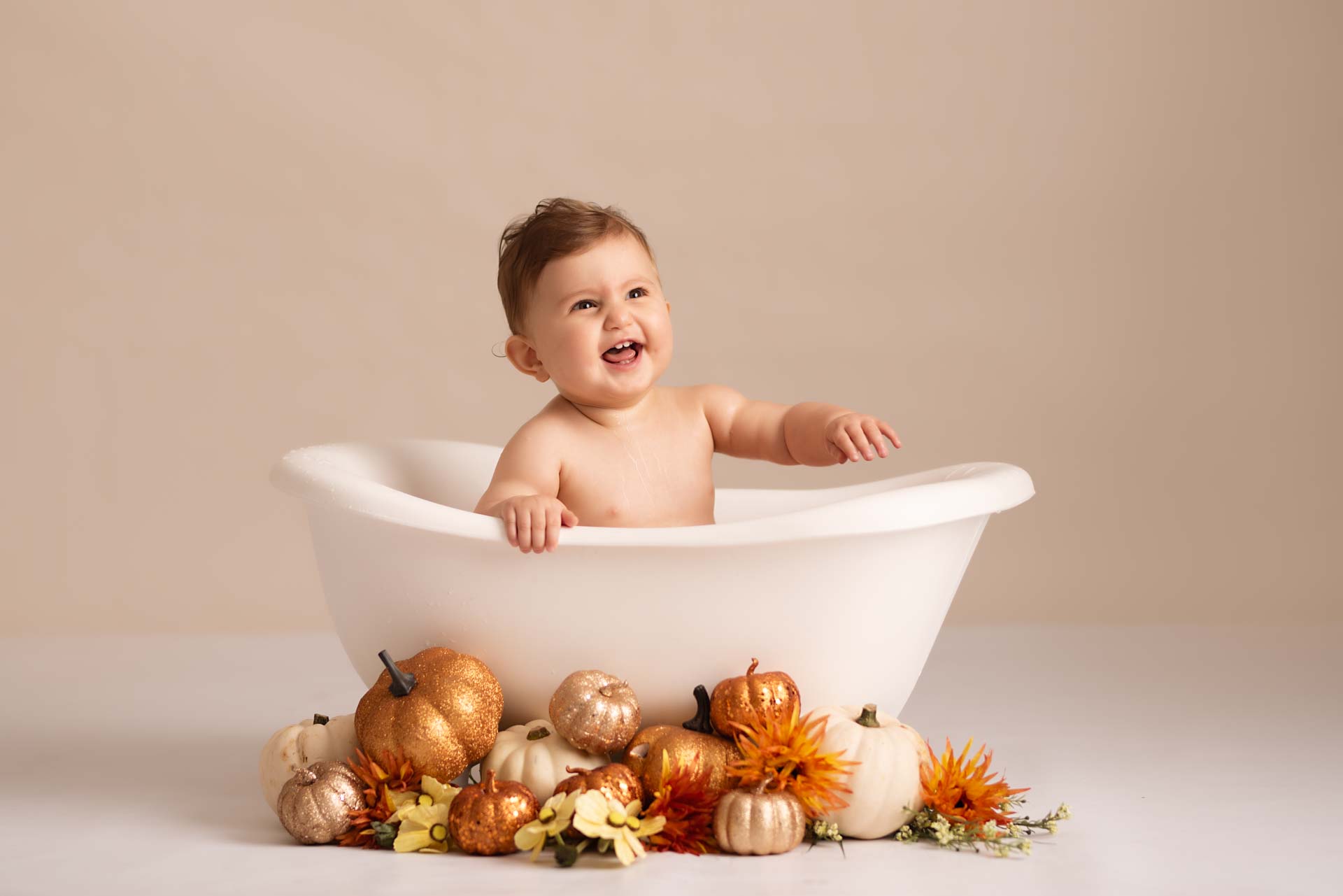 One year old sitting in a roll top bath surrounded by different coloured pumpkins