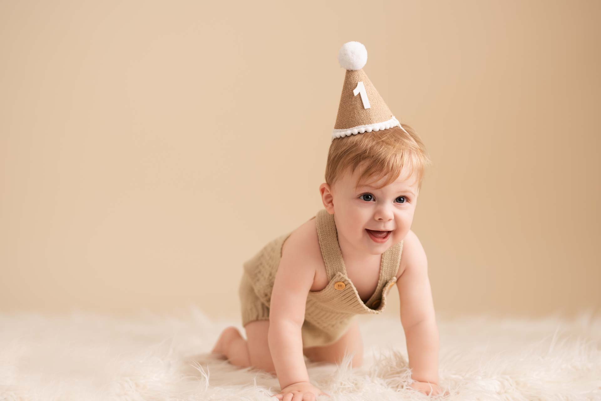 One year old boy wearing a brown party hap crawls towards the camera with a big smile on his face