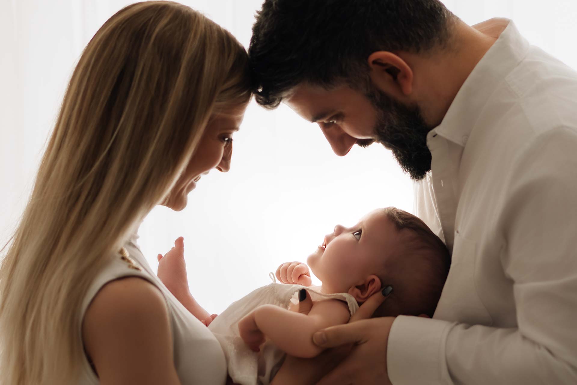 Mother and father face to face with their foreheads touching have their baby girl in between them looking up at them