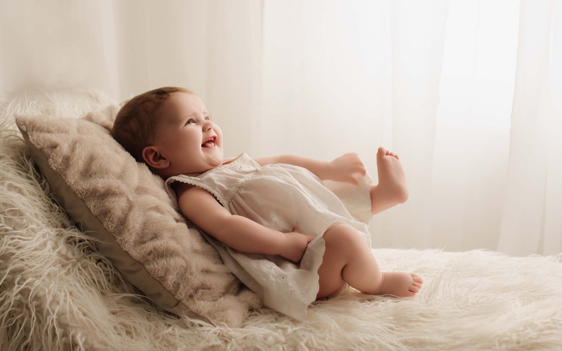 5 month old girl smiling and laughing leaning on a cushion on top of a white fluffy blanket