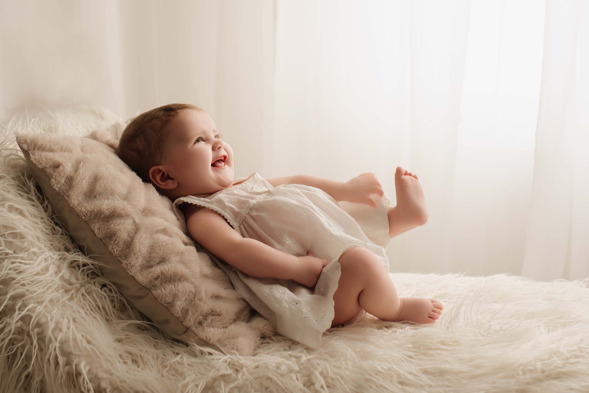 5 month old girl smiling and laughing leaning on a cushion on top of a white fluffy blanket