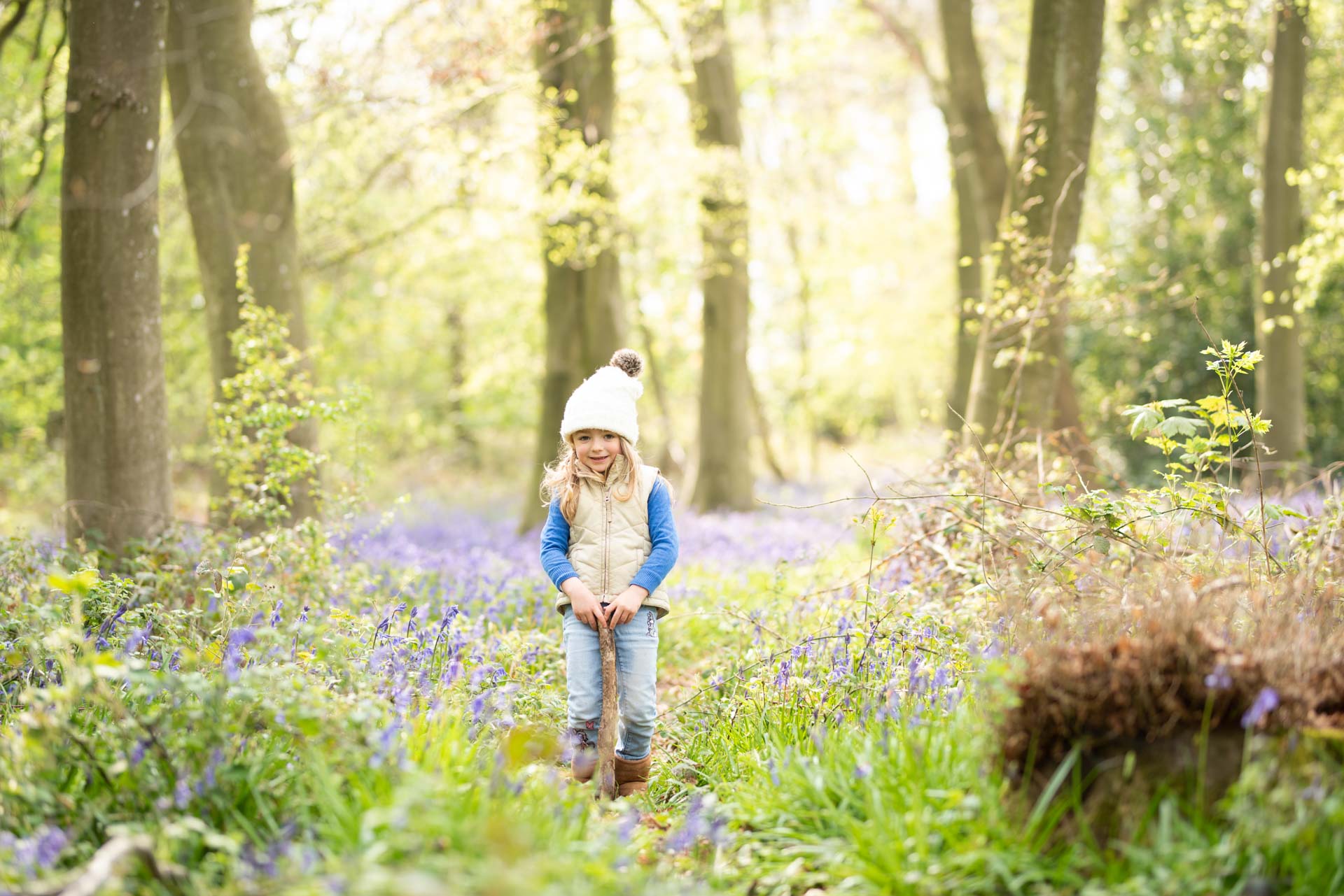 Toddler wearing blue jeans and a beige gilet standing in a bluebell woodland holding a large stick which she has found.