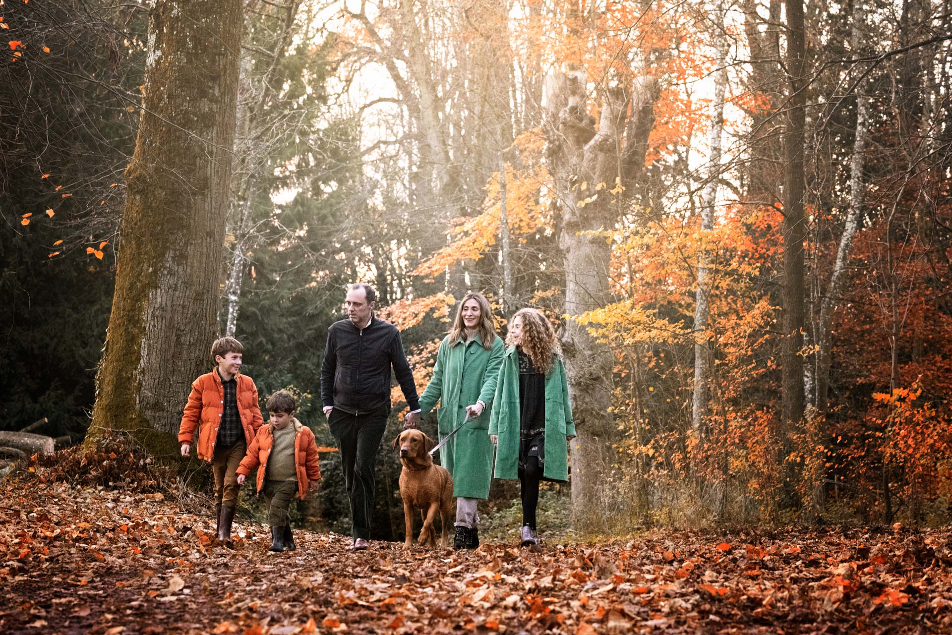 A family of five taking their dog for a walk through the woods in the autumn. The boys are wearing orange and the girls are wearing green