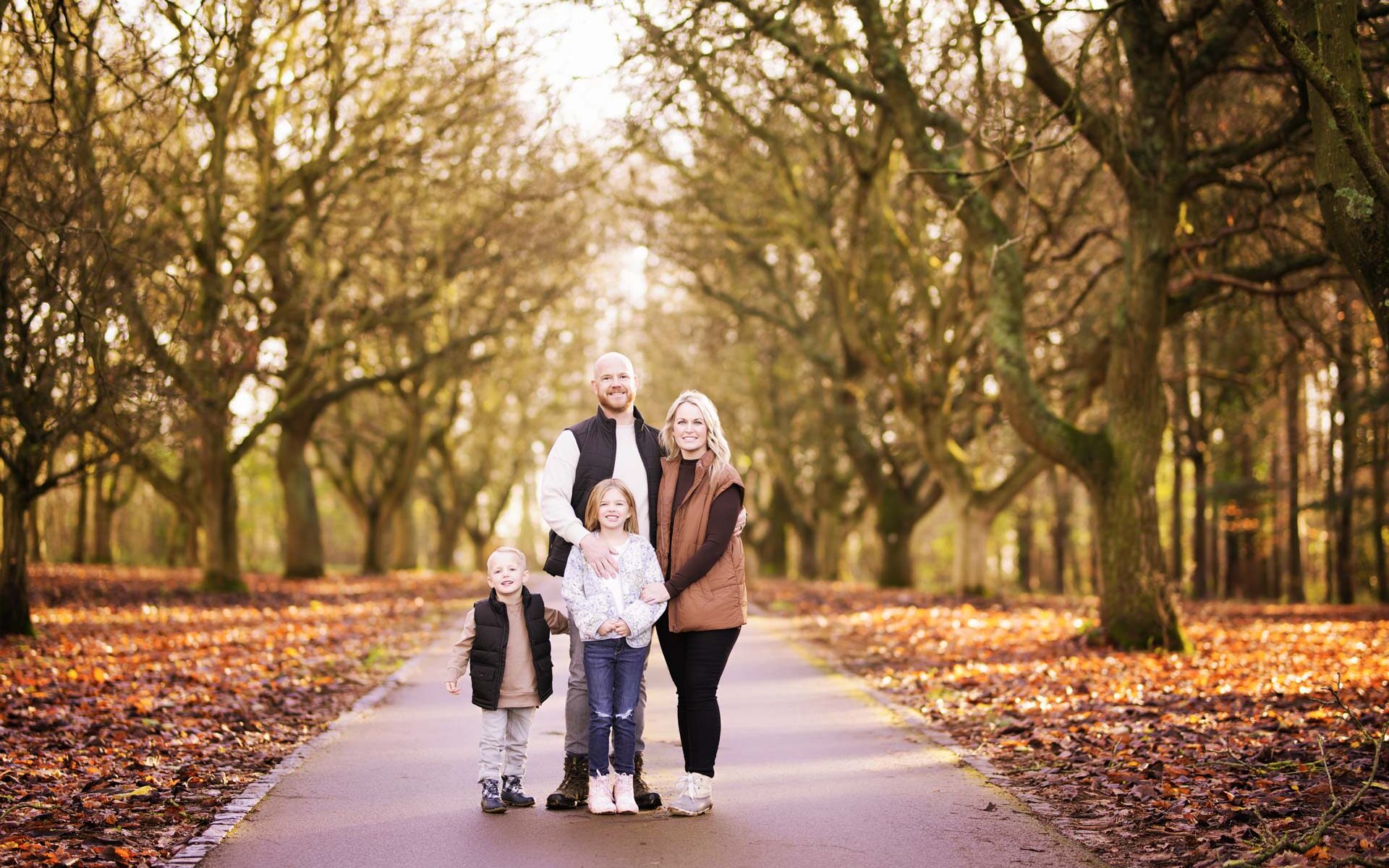 Family of four stand on a tarmac road lined by trees as the autumn light begins to fade