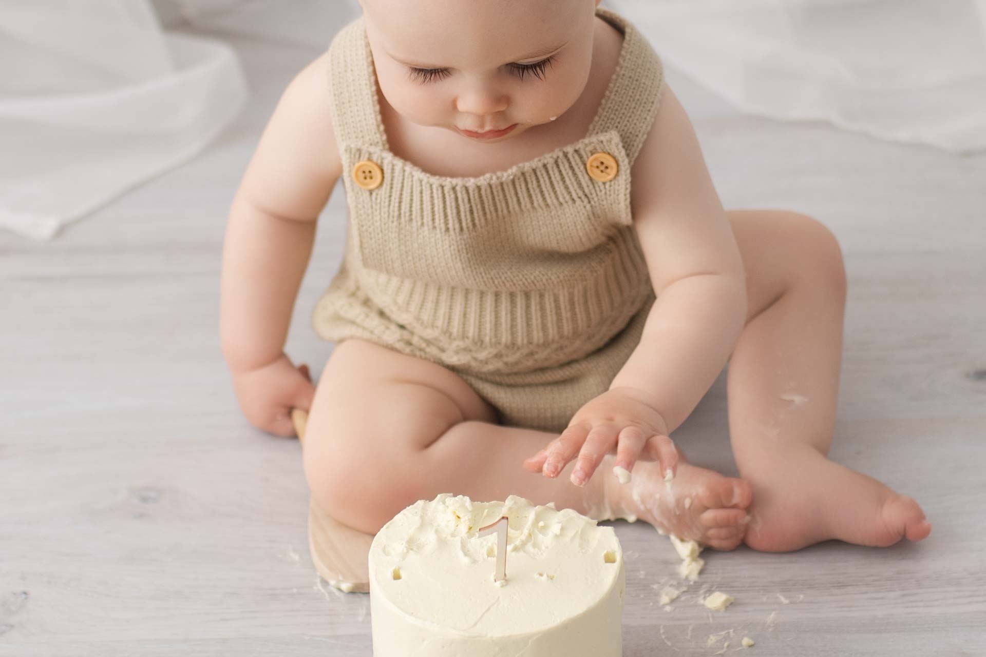 A one year old tentatively touches the icing on his first birthday cake. Some of it is on his foot
