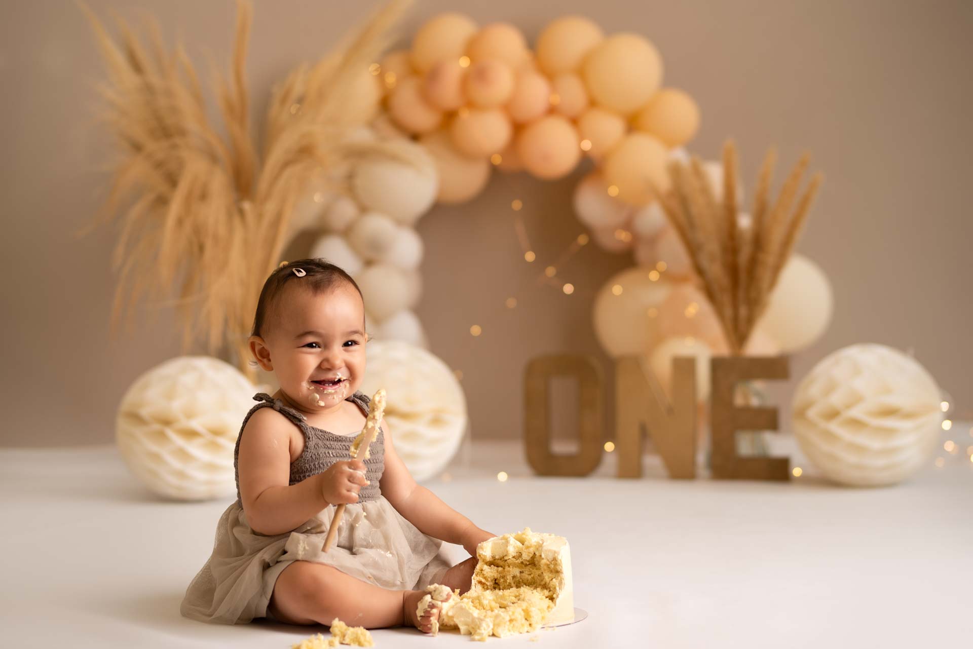 Baby girl holding a wooden spoon. Her face is covered in icing and next to her is a half smashed cake.