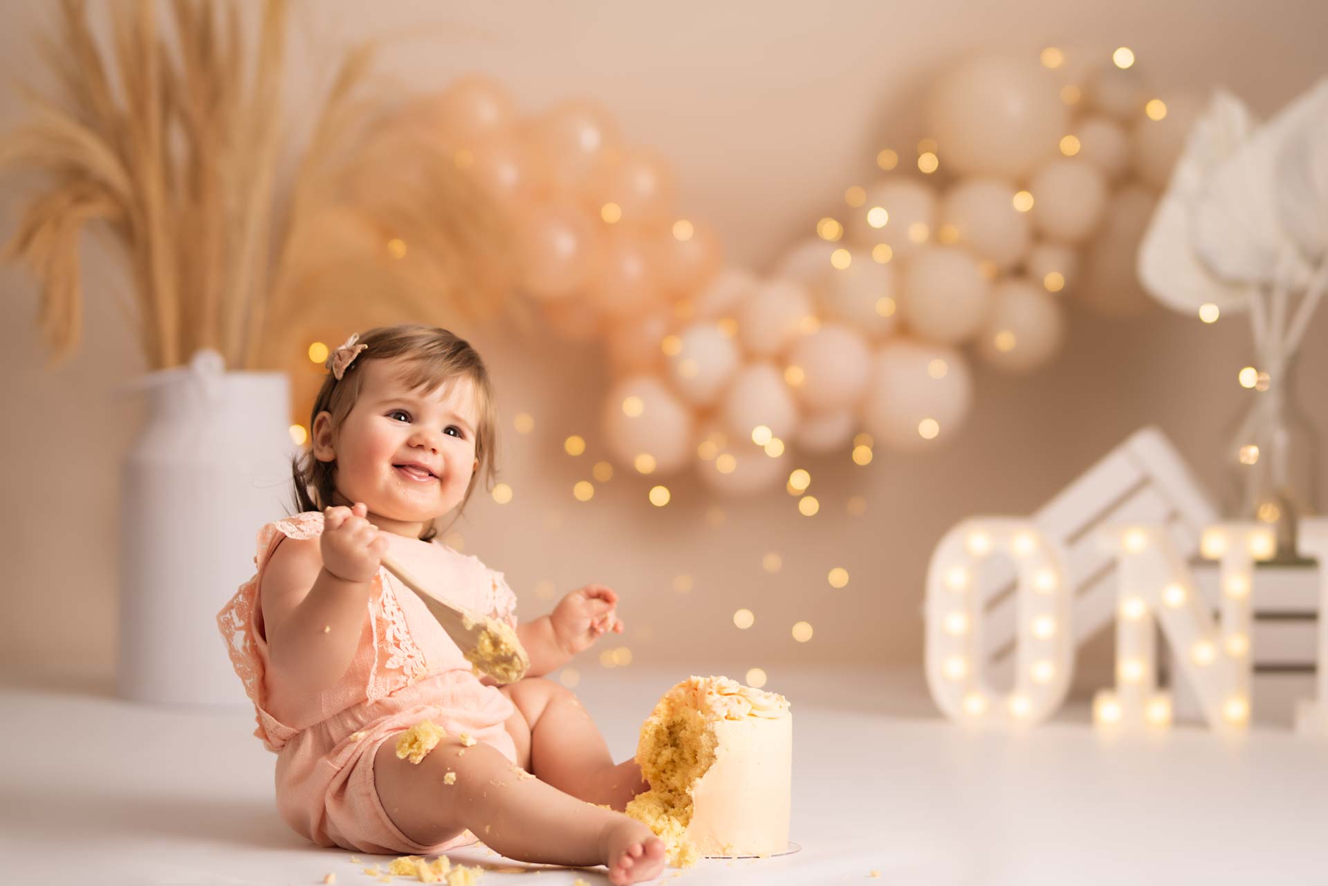 First birthday girl holding a wooden spoon and eating her birthday cake. The colour scheme is peach