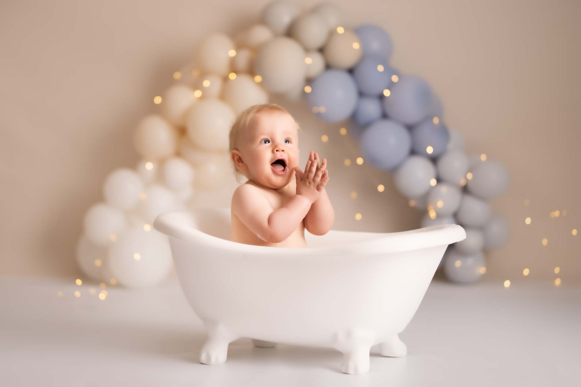 Baby boy sitting in a white plastic bath clapping his hands. In the background are white and blue balloons of different shades