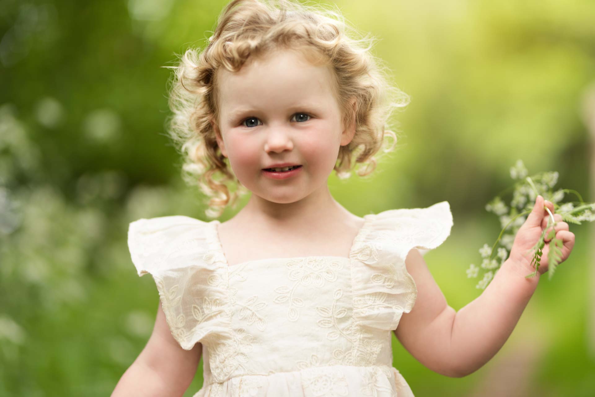 3 year old girl with curly hair holding some flowers that she has picked on her walk in the countryside. She is wearing a pale yellow dress.