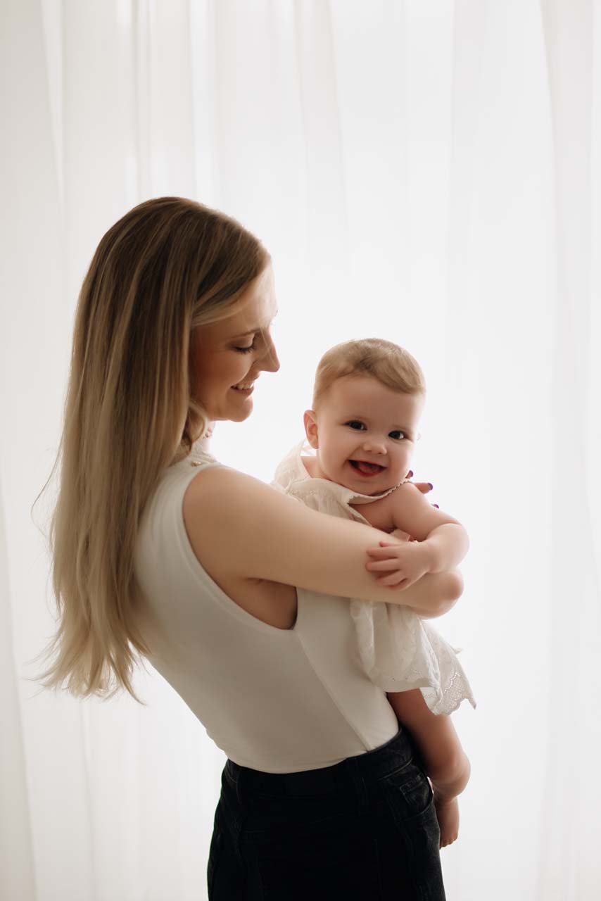 Baby girl being cuddled by her mother and giving the camera a cheeky smiling. The mother is standing by a window with net curtains