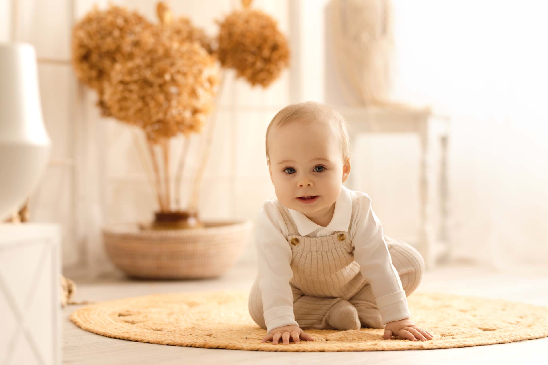 Young boy sitting on a mat wearing beige romper and white top looking at the camera