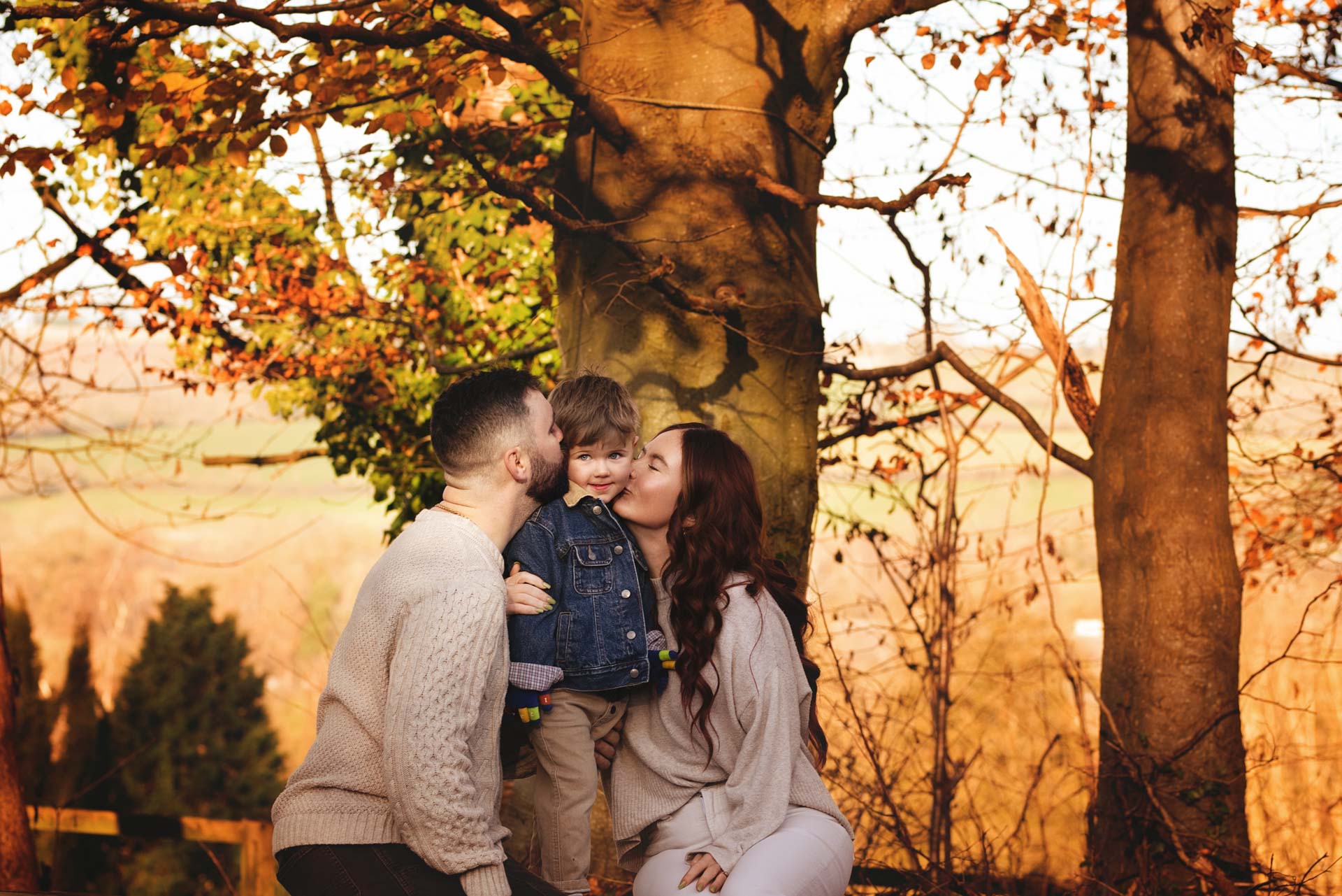 A couple both kissing their son on the cheek. They are standing in front of a tree surrounded by autumn colours