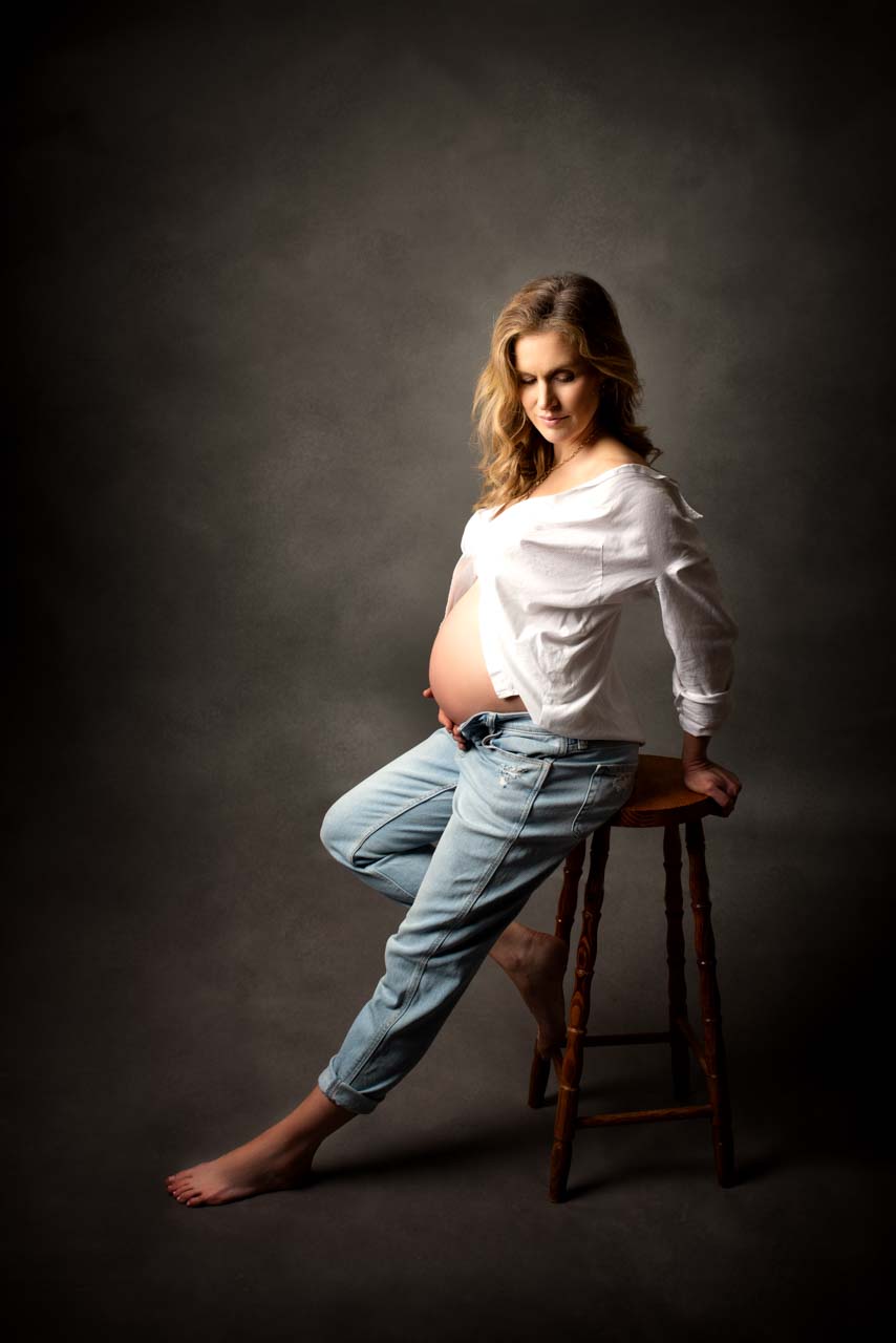 Mum-to-be sitting on the edge of a stool holding her bump. She is wearing jeans and I white shirt.