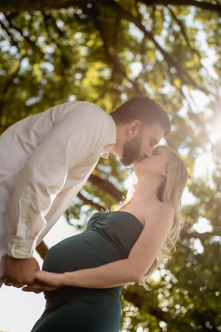Pregnant couple kissing and holding hands. She is wearing a dark green dress. He is wearing a smart white shirt.