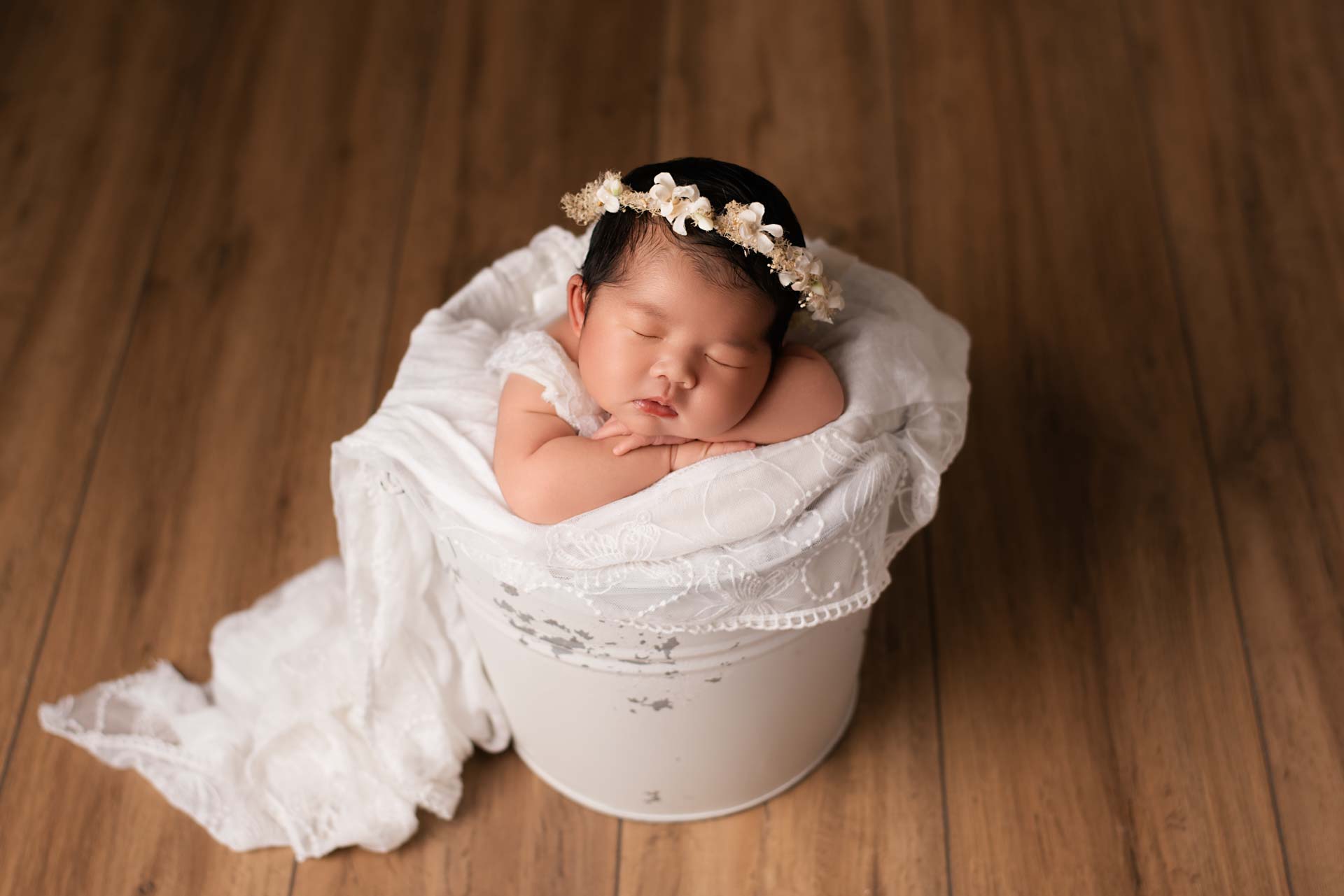 Newborn baby girl asleep in a white bucket on brown wooden floorboards