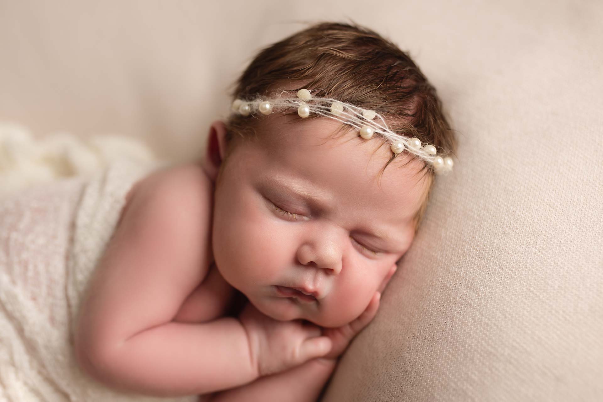 Newborn baby with her hands under her chubby cheeks wearing a pearl headband