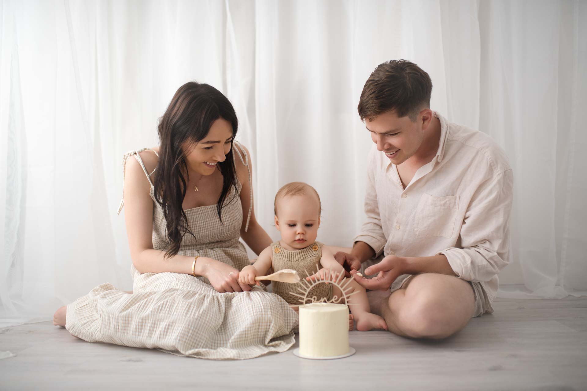 A mummy and daddy sit next to their baby boy at his first birthday photoshoot. He is about to hit a cake with a wooden spoon