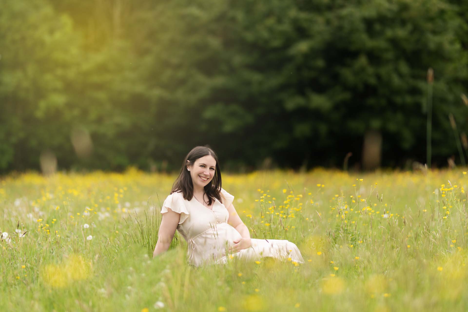 Expectant mum wearing a cream dress sitting in a field of wild flowers.