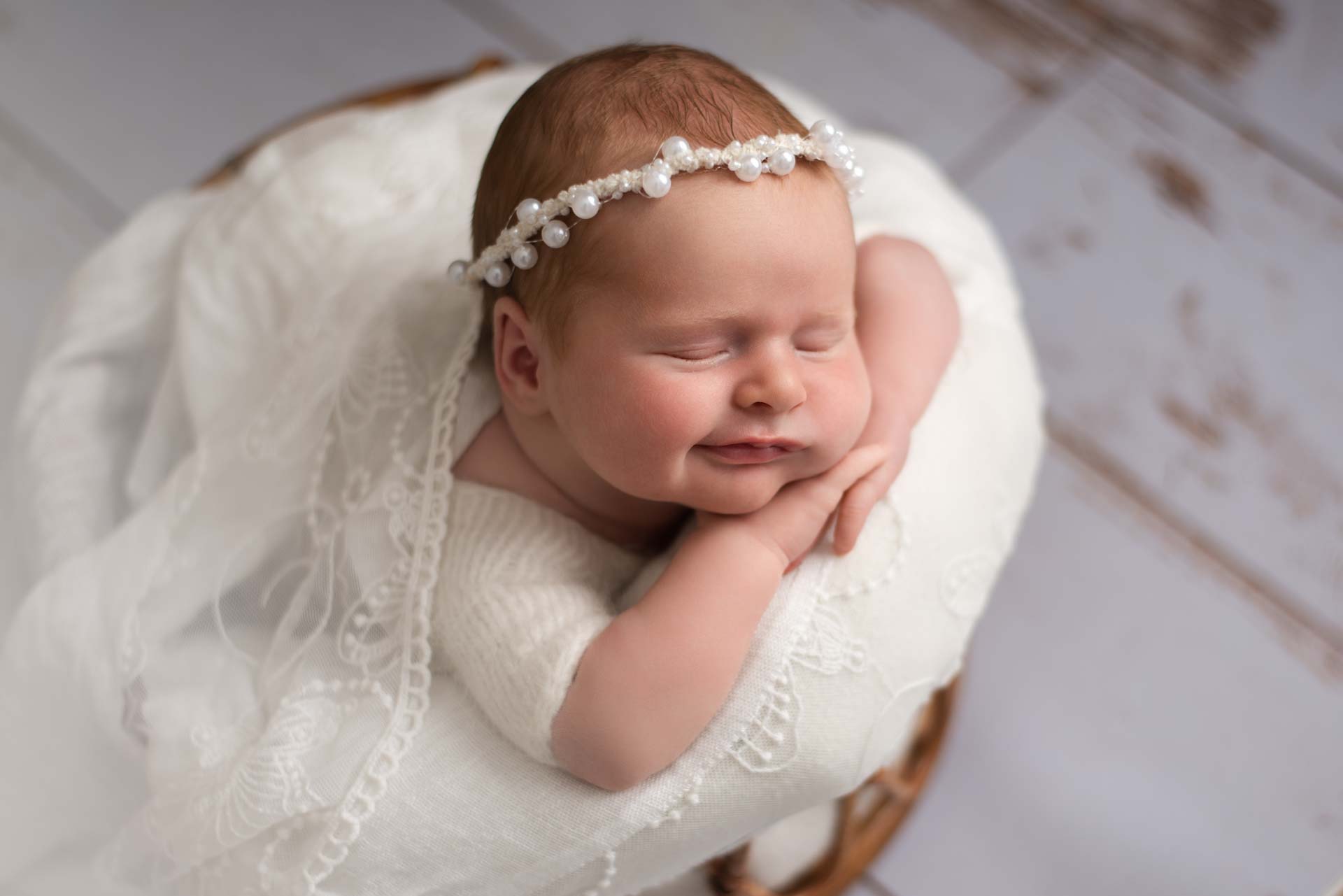 Smiling baby girl lying on her front in a bamboo basket. The colour scheme is all white including the pearl headband and floorboards