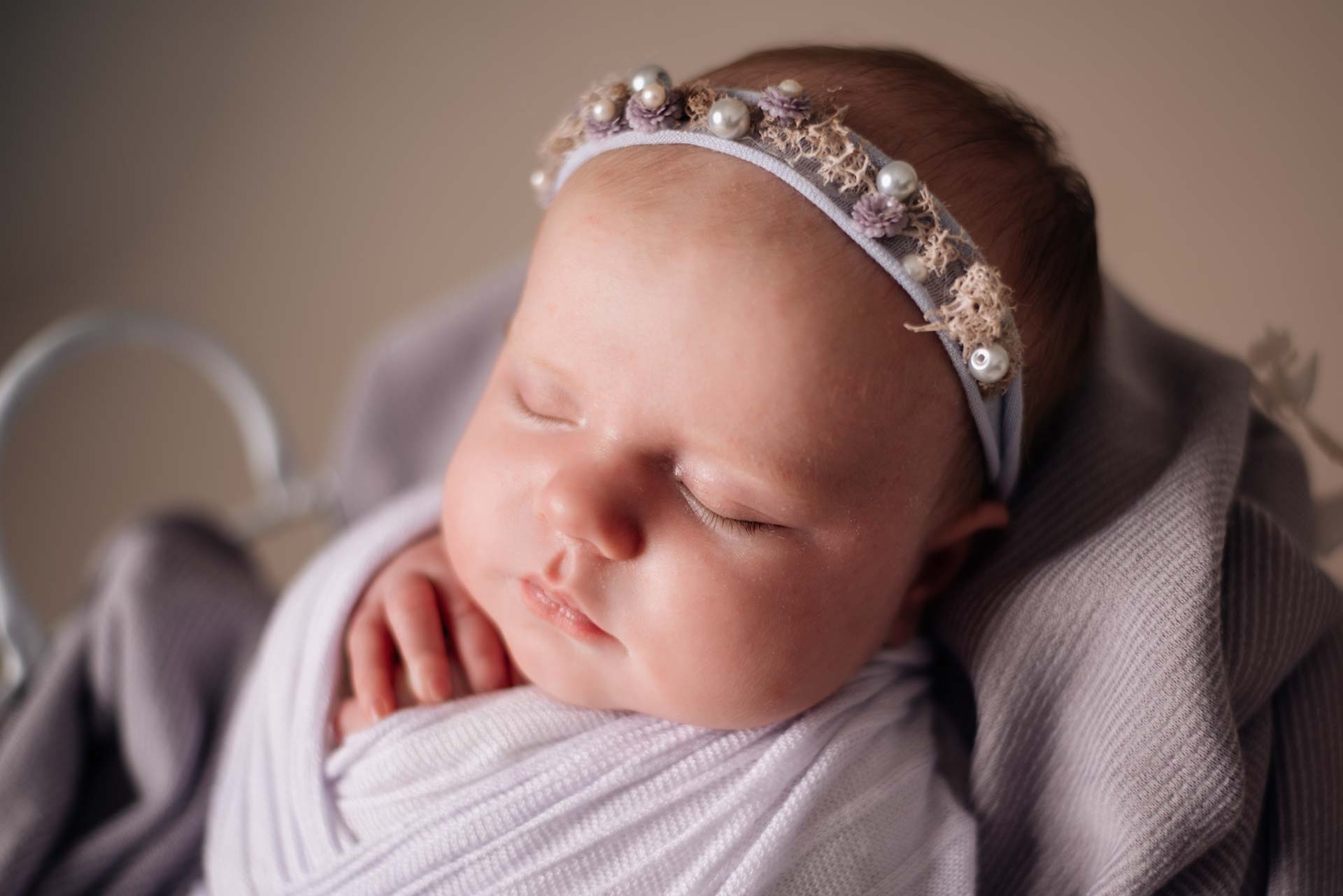 Swaddled baby girl in a white metal basket. The wrap, blanket and headband are all lilac