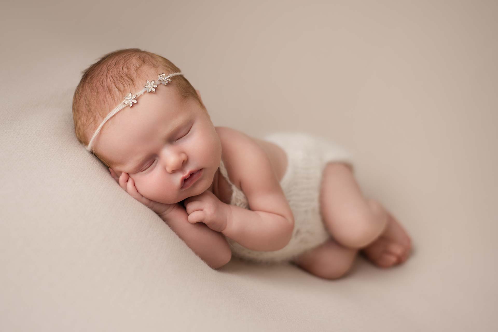 Newborn baby sleeping on her side on the photographers bean bag wearing a white romper and a delicate headband with three metal flowers on it