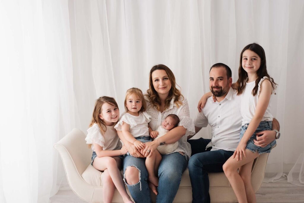 Family of six pose on a chaise for their family photoshoot. The background is white net curtain