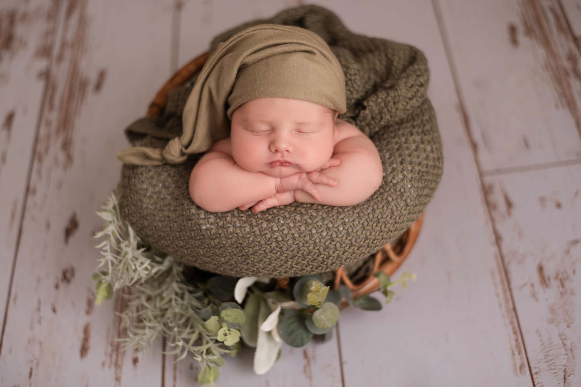Baby boy lying on his front in a bamboo baskets. The colour scheme is sage green and the basket is placed on white wooden floorboards