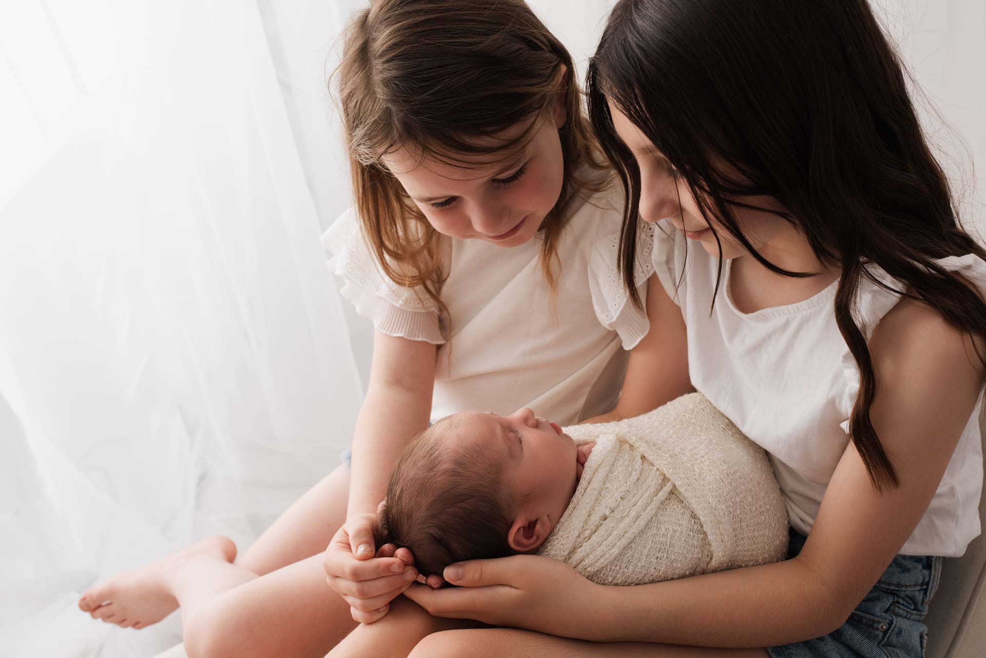Two sisters holding their new baby sister who is swaddled