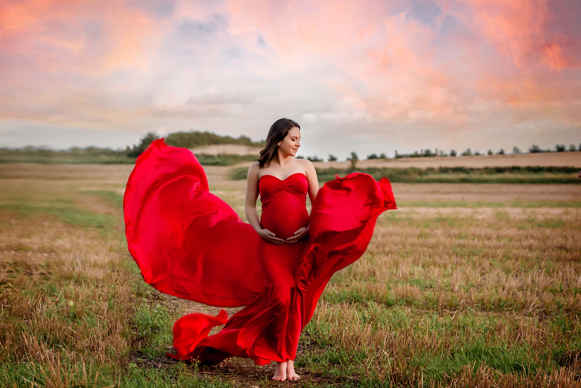Pregnant women wearing red dress standing in a field at sunset. The dress is blowing in the wind.