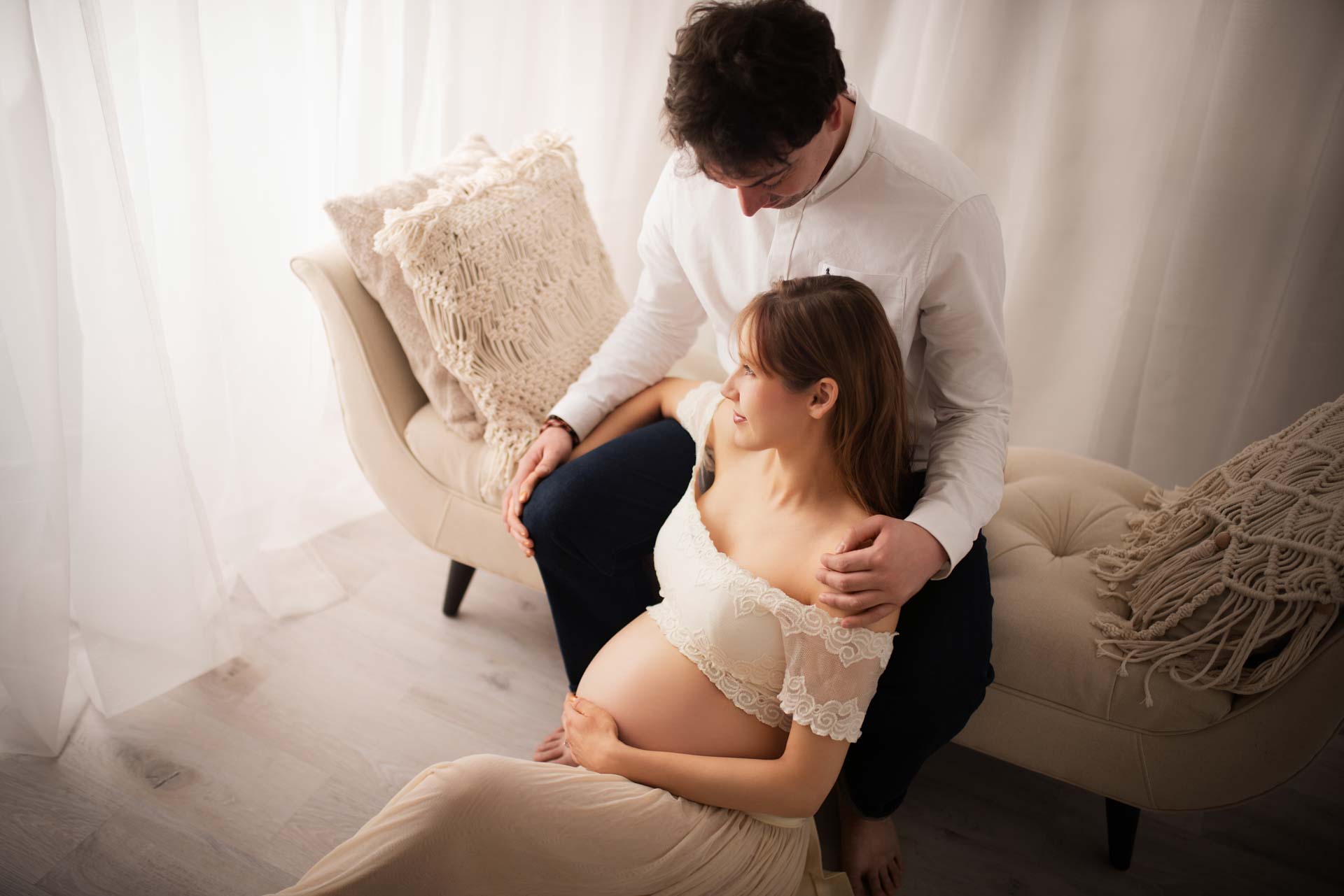 Pregnant lady sitting on the floor wearing a lace top and flowing dress. Her partner sitting on a chaise supporting her.